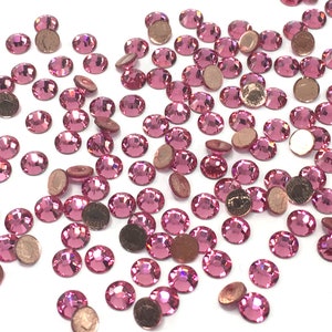 May include: A large quantity of pink rhinestones, also known as crystals or gems, scattered on a white surface. The rhinestones have a faceted design and a silver backing.