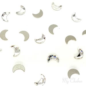 May include: A collection of clear crystal crescent moon shaped nail art embellishments. The rhinestones are scattered on a white background.