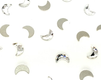 Puede incluir: Una colección de adornos para uñas de cristal transparente en forma de media luna. Los diamantes de imitación están esparcidos sobre un fondo blanco.