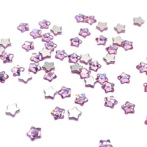 May include: A collection of star-shaped embellishments. Some are silver, others are light pink with a faceted, gem-like appearance. The stars are scattered on a white surface, creating a sparkling effect.