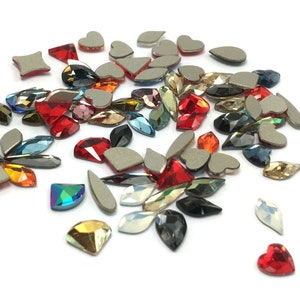 May include: Assortment of colorful rhinestones in various shapes, including hearts, teardrops, and ovals. The rhinestones are scattered on a white surface.