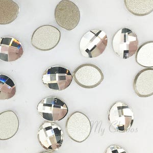 May include: Close-up of oval-shaped rhinestones with a faceted, clear crystal design. Some rhinestones have a silver backing, while others are clear. The rhinestones are scattered on a white surface, reflecting light.