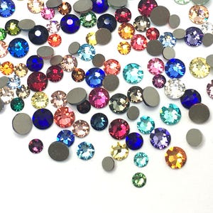 May include: A selection of multi-coloured rhinestones in different sizes and hues, including blue, pink, green, yellow, and clear. Some rhinestones have a flat, grey backing. The gemstones are scattered on a white surface.