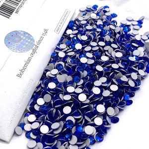 May include: A large pile of royal blue self-adhesive rhinestones. The rhinestones are scattered on a white surface. The rhinestones are packaged in a white box with the text "Bohemian crafted since 1948" and "Made in the Czech Republic".