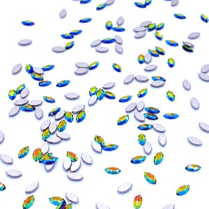 Può includere: Una collezione di piccoli cristalli a forma di marquise sparsi su una superficie bianca. Ogni pezzo presenta una parte superiore sfaccettata con una finitura iridescente blu, verde e gialla, mentre il retro è una base metallica argentata piatta.