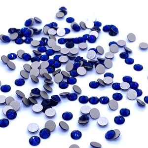 Op de afbeelding: Een spreiding van gefacetteerde, diepblauwe strass-steentjes met zilveren achterkant. De edelstenen zijn verspreid over een wit oppervlak, met verschillende maten en vormen. De strass-steentjes zijn reflecterend en vangen het licht.