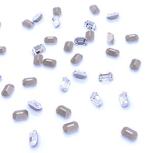 May include: A collection of small, rectangular, faceted crystals scattered across a white surface. The crystals alternate between clear, transparent glass-like pieces and opaque, matte-finished beige or light tan colored stones.
