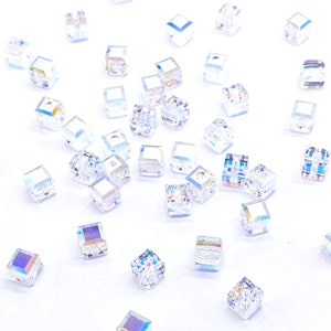 May include: A scattering of iridescent clear glass cube beads on a white background. The beads are small and have a faceted design.