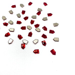May include: Scattered on a white surface are numerous red and silver-colored gemstone embellishments. The red gems are rectangular with a pointed end, while the silver gems are geometric. These decorative elements are suitable for crafts.