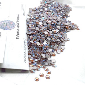 Puede incluir: Primer plano de un montón de piedras de cristal de color rosa claro iridiscente sobre un fondo blanco. Las piedras de cristal están en una bolsa pequeña con el texto "PRECiosa COMPONENTS Bohemian crafted since 1948." impreso en el lateral.