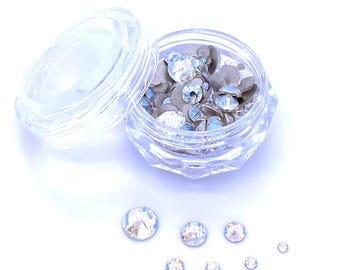 May include: A clear, faceted plastic jar with an open lid, filled with silver and clear rhinestones. Several loose rhinestones of varying sizes are scattered around the jar on a white surface. The rhinestones have a sparkling, iridescent quality.