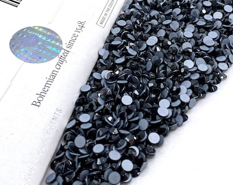 May include: A close-up of a pile of black rhinestones. The rhinestones are small and round, with a flat back and a pointed top. The rhinestones are arranged in a random pattern, and they are all the same size. The rhinestones are on a white background. The text on the box reads "Bohemian crafted since 1948. Made in the Czech Republic."