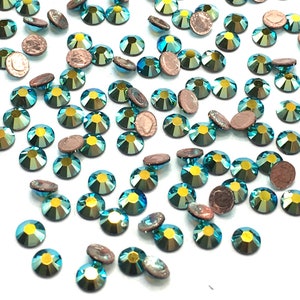 Puede incluir: Una dispersión de piedras de cristal azul turquesa con un acabado iridiscente dorado. Las piedras de cristal están dispersas sobre un fondo blanco.