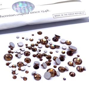 May include: A scattering of brown iridescent rhinestones, some with adhesive backing, on a white surface.  The rhinestones are various sizes and shapes.