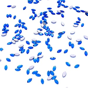 Può includere: Una collezione di piccoli strass blu a forma di marquise sparsi su una superficie bianca. Ogni singolo cristallo presenta una silhouette appuntita a forma di occhio con un colore blu vibrante e saturo e un retro piatto dai toni metallici.