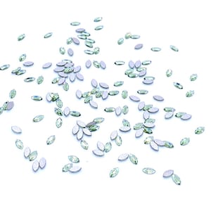 May include: Scattered on a white surface are numerous light green, oval-shaped rhinestones. Each rhinestone has a silver backing, creating a sparkling effect. The rhinestones are ideal for embellishing crafts, clothing, or accessories.