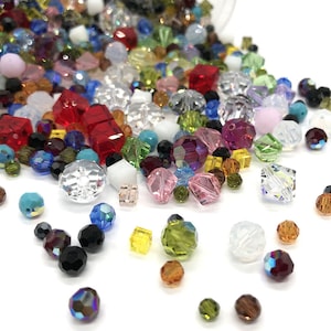 Peut inclure: Un grand assortiment de perles de verre colorées de différentes formes et tailles, notamment rondes, carrées et facettées. Les perles sont éparpillées sur une surface blanche.