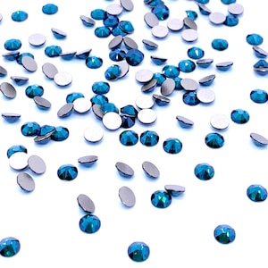 Può includere: Una collezione di piccoli cristalli rotondi sfaccettati di colore verde acqua sparsi su una superficie piana. Ogni cristallo presenta una parte superiore a taglio brillante con molteplici superfici riflettenti e un retro metallico piatto e opaco.
