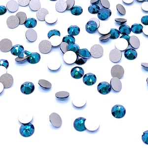 Può includere: Una collezione di piccoli cristalli rotondi e sfaccettati color verde acqua sparsi su una superficie bianca. Ogni cristallo presenta una superficie superiore brillante e sfaccettata che riflette la luce, con un retro metallico opaco visibile.