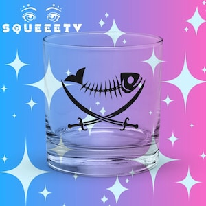 Puede incluir: Un vaso transparente con un diseño negro de temática pirata. El diseño presenta una espina de pescado, espadas cruzadas y la palabra "SQUEEETV" en blanco. El vaso tiene forma cilíndrica recta.
