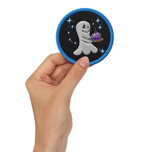 Puede incluir: Parche bordado con un fantasma sonriente sosteniendo un pastel de cumpleaños morado en una bandeja dorada. El parche tiene un fondo negro con estrellas azules y un borde azul. El fantasma es blanco.