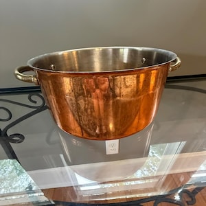 May include: A copper-colored metal bucket with two brass handles. The interior of the bucket is a lighter, silver color. The bucket is sitting on a glass table with a decorative black metal base.