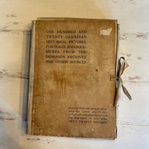 May include: A vintage book with a tan cover and a ribbon tie. The cover features text that reads "ONE HUNDRED AND TWENTY CANADIAN HISTORICAL PICTURES, PORTRAITS, AND DOCUMENTS." The book appears aged with some staining.