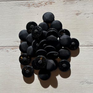 May include: A pile of black, fabric-covered buttons with a shiny, dark centre. The buttons are scattered on a light-coloured, wooden surface. The buttons have a classic design, suitable for various sewing projects.