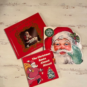 May include: Three vintage Christmas books are displayed on a white wooden surface. The books are red and feature illustrations of Santa Claus. One book is titled "A Special Place for Santa," another is "The Christmas Santa Almost Missed," and the third is "The Santa Claus Book."