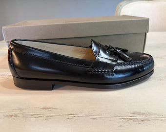 May include: A pair of black leather penny loafers with a tassel detail. The shoes are displayed in front of a cardboard box on a light-colored wooden surface. The loafers have a classic, polished appearance.