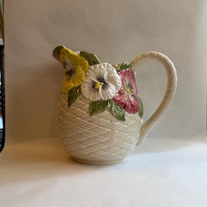 May include: Cream-colored ceramic pitcher with a woven basket design. The pitcher is adorned with colorful pansy flowers in yellow, white, and pink, with green leaves. The handle is a twisted rope design.