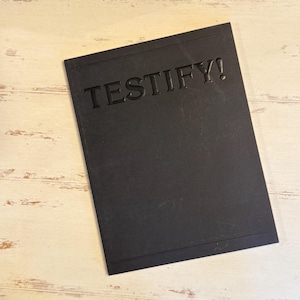 Pode incluir: Uma placa retangular preta com a palavra "TESTIFY!" em relevo na superfície. A placa está sobre um fundo de madeira clara e desgastada. As bordas da placa têm uma borda sutil.