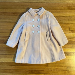 May include: A light pink wool coat with a double-breasted design. The coat features a rounded collar, two front pockets, and six large, white buttons. The coat is designed for children.
