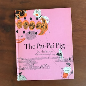 May include: A hardcover book titled "The Pai-Pai Pig" by Joy Anderson, with illustrations by Jay Yang. The cover is pink with a whimsical illustration of a butterfly and other abstract shapes. A child is flying a kite on the cover.