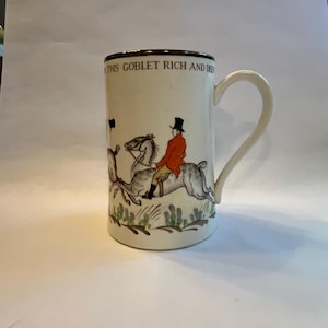 Könnte beinhalten: Cremefarbene Keramiktasse mit braunem Rand und Henkel. Sie zeigt eine Jagdszene mit einem Reiter in einem orangefarbenen Mantel auf einem Pferd und dem Text "THIS GOBLET RICH AND DEEP".