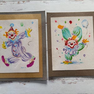Puede incluir: Dos pinturas en acuarela enmarcadas de payasos. Uno lleva un atuendo morado con un sombrero rojo y blanco, y el otro un atuendo verde con una corona. Ambas pinturas tienen globos de colores.