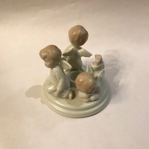 May include: A ceramic angel figurine with three cherubic figures arranged around a central candle holder. The angels are in a kneeling or seated position, with wings and light brown hair. The base is a light gray color.