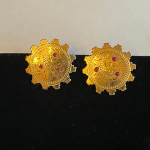 Könnte beinhalten: Ein Paar goldfarbene Ohrstecker. Jeder Ohrring hat ein zahnradähnliches Design mit einem floralen Muster und drei kleinen roten Edelsteinen. Die Ohrringe werden auf einer schwarzen Oberfläche präsentiert.