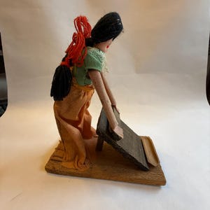 May include: A handmade doll depicting a woman in traditional attire. The doll wears a green blouse, orange skirt, and has black hair with red accents. She is positioned as if working with a wooden tool on a wooden base.