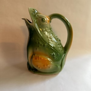 May include: A ceramic pitcher in the shape of a frog. The pitcher is primarily green with orange accents on the belly and handle. The frog's mouth is open, forming the spout. The pitcher has a textured surface with small bumps.