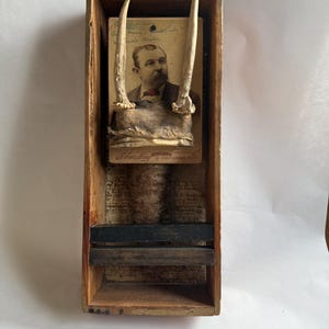 Może przedstawiać: Drewniane pudełko w stylu vintage z portretem mężczyzny, ozdobione porożem jelenia. Portret umieszczono na tle postarzanego papieru i odręcznego pisma. Pudełko ma rustykalny, postarzany wygląd.