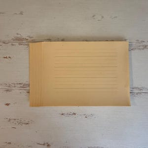 Può includere: Una pila di fogli di carta a righe color beige. La carta ha linee orizzontali e sembra vintage o invecchiata. La carta è impilata su una superficie di legno chiaro e usurato.