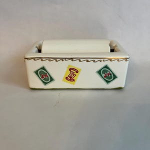 Puede incluir: Un soporte rectangular de cerámica blanquecina para rodillo quitapelusas con ribete dorado y diseño de ondas. Presenta tres gráficos tipo sello con el texto "TODAY VALUE". El rodillo es blanco y se encuentra dentro.