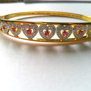 May include: A gold-tone hinged bangle bracelet featuring a repeating pattern of heart-shaped links. Each heart is encrusted with small clear stones and centers a single red stone. The inner band is engraved with the text I loved you yesterday, I love you today, I love you more each passing day.