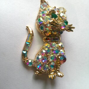 May include: A gold-toned cat brooch featuring a textured metal body encrusted with iridescent, multi-colored rhinestones. The cat has green rhinestone eyes, a collar with rectangular stones, and a tail accented with rectangular iridescent crystals.