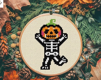 May include: A cross stitch pattern featuring a skeleton dressed as a pumpkin. The pattern is in a circular embroidery hoop with a white background. The skeleton is wearing a black and white skeleton costume with a pumpkin head. The pumpkin has a black face with a white smile and eyes. The pumpkin is orange with a green stem.