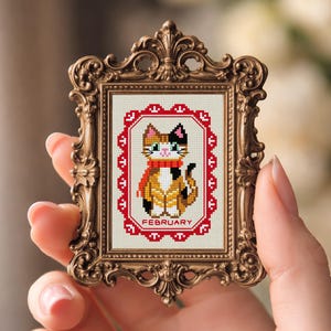 May include: A framed cross-stitch featuring a calico cat wearing a red scarf, with the month "February" in a red border. The frame is ornate gold-colored metal. The text "Dona Stitch" and "Donastitch.etsy.com" are visible.