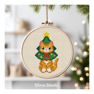 May include: A cross-stitch embroidery hoop featuring a ginger cat wearing a green Christmas tree costume, topped with a yellow star. The cat has white whiskers and is set against a beige background. The text "Dona Stitch" is at the bottom.