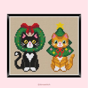 以下が含まれることがあります： 2匹の猫を描いたクロスステッチアート。1匹はクリスマスのリースをつけた黒猫、もう1匹はクリスマスツリーのコスチュームを着たオレンジ色の猫です。どちらも白いひげとハート型の模様があります。アートワークは黒いフレームに入っています。