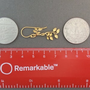 May include: Gold leaf earrings with a delicate branch design. The earrings are approximately 2.5 cm long and are shown next to a ruler and two coins for size reference.
